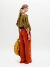 Load image into Gallery viewer, A woman stands sideways against a plain white background, wearing the Daia Blouse in olive green with a V-cut neckline, paired with loose orange pants and holding a textured yellow bag.
