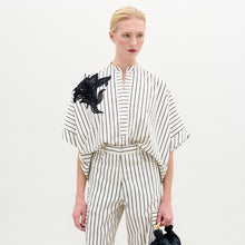 Load image into Gallery viewer, A person with blond hair in a low ponytail wears the Daia Blouse, a loose, white 100% cotton top with black vertical stripes and short sleeves, standing against a plain light background.
