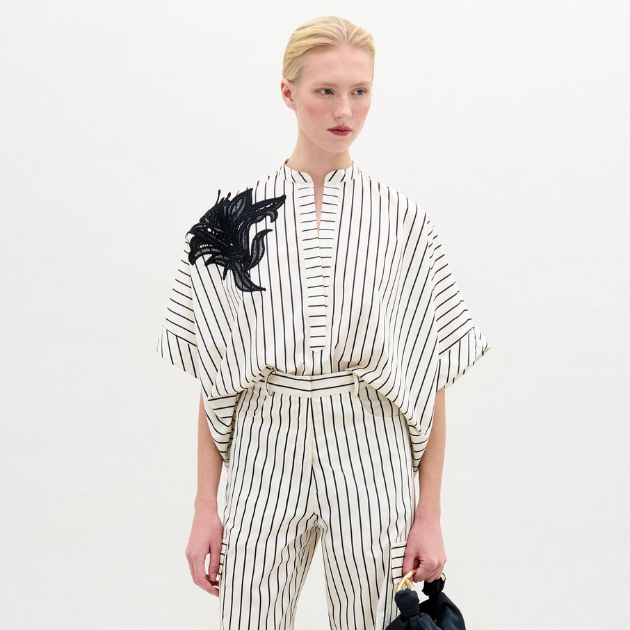 A person with blond hair in a low ponytail wears the Daia Blouse, a loose, white 100% cotton top with black vertical stripes and short sleeves, standing against a plain light background.