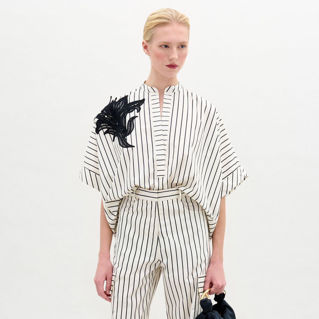 A person with blond hair in a low ponytail wears the Daia Blouse, a loose, white 100% cotton top with black vertical stripes and short sleeves, standing against a plain light background.