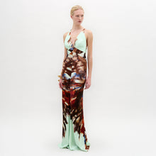 Load image into Gallery viewer, A woman stands against a plain white background wearing the Daniella Dress—a sleeveless maxi with a deep V-neckline, featuring a light blue top and a brown and red abstract print on the lower half.
