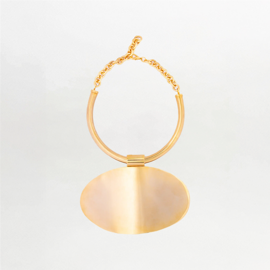 The Danielle Necklace features a gold-tone choker with a thick, round chain and a large flat oval pendant at the center, creating a minimalistic silhouette against a plain white background.