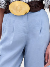 Load image into Gallery viewer, A woman stands against a plain background wearing a striped blouse with blue floral appliqué, the Darnell Pant in light blue, a brown belt with a large buckle, gold sandals, and holding a beige bag.
