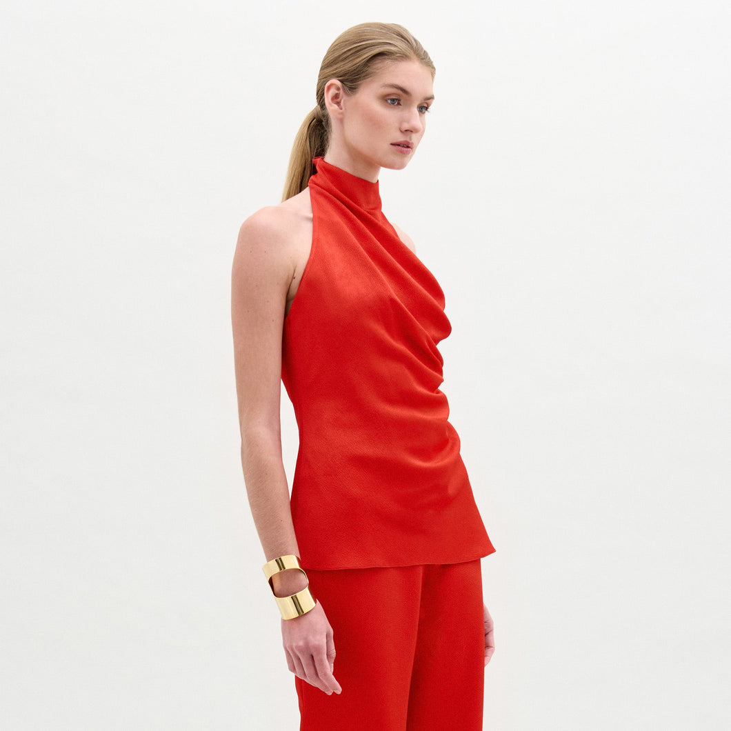 A woman stands against a plain white background wearing the lustrous hammered satin Dina Blouse, a high-neck sleeveless top, with matching red pants. Her hair is tied back and she accessorizes with a wide gold bracelet.