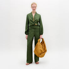 Load image into Gallery viewer, A woman stands against a plain background wearing wide-leg Emine Pants paired with a green Italian fabric blouse, holding a large textured mustard-yellow bag.
