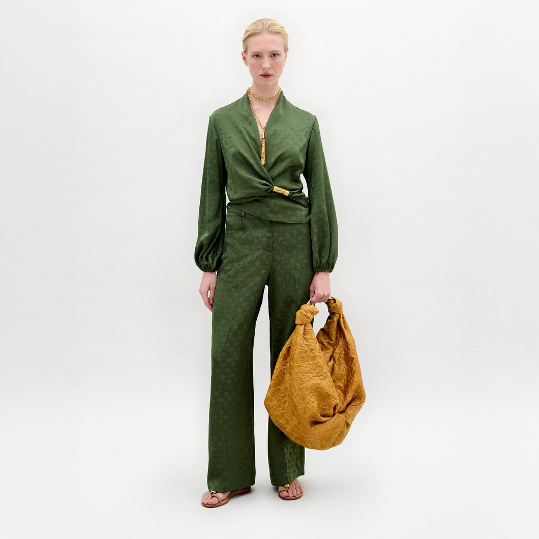 A woman stands against a plain background wearing wide-leg Emine Pants paired with a green Italian fabric blouse, holding a large textured mustard-yellow bag.