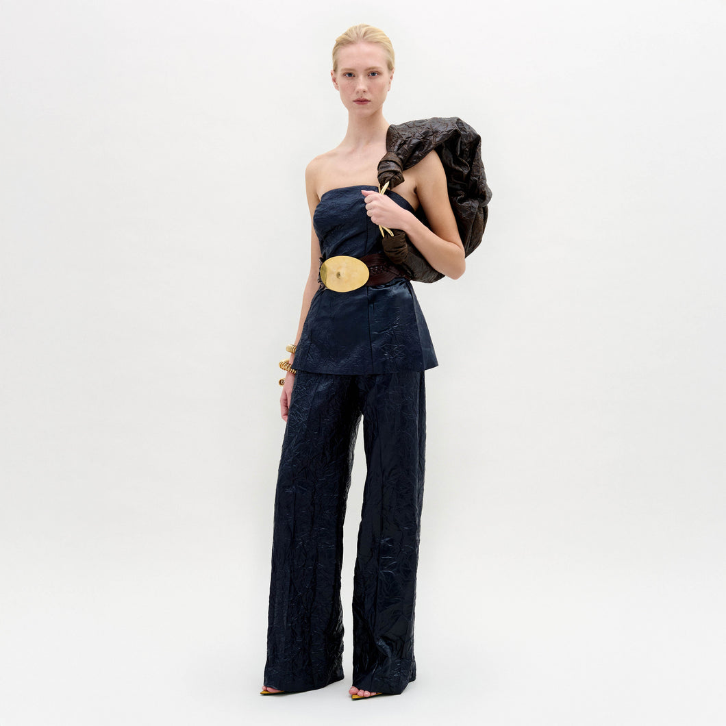 A woman stands against a white background wearing the Emine Pant—a high-waisted, wide-leg silhouette—paired with a dark strapless top, gold belt, bangles, and a textured bag slung over her shoulder.
