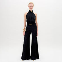 Load image into Gallery viewer, A person stands against a white background in the Enara Jumpsuit, a black halter-neck piece made from luxurious Italian fabric with wide-leg pants, accessorized with elegant gold bangles.
