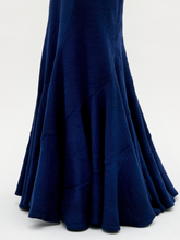 Load image into Gallery viewer, A woman wearing the Florina Skirt—a floor-length navy blue skirt with a mermaid silhouette and gathered knot detail at the waist—paired with gold jewelry, stands against a plain white background.
