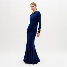 Load image into Gallery viewer, A woman wearing the Florina Skirt—a floor-length navy blue skirt with a mermaid silhouette and gathered knot detail at the waist—paired with gold jewelry, stands against a plain white background.
