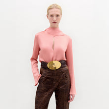 Load image into Gallery viewer, A woman stands against a plain background wearing the Gaby Blouse, a pink high-neck top with a front slit, paired with brown textured pants and a wide belt featuring a large gold oval buckle.
