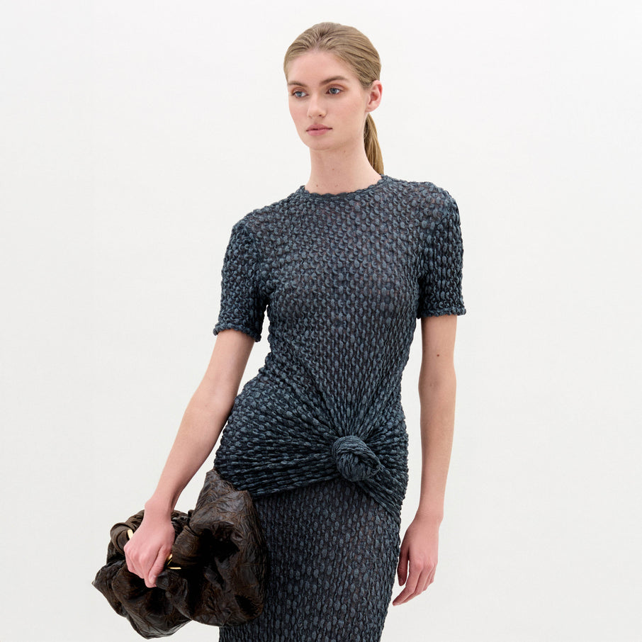 A woman with blonde hair in a low ponytail wears the Gen Tshirt, a textured, fitted, dark short-sleeve top with knot detail, and holds a large dark ruched bag against a plain white background.