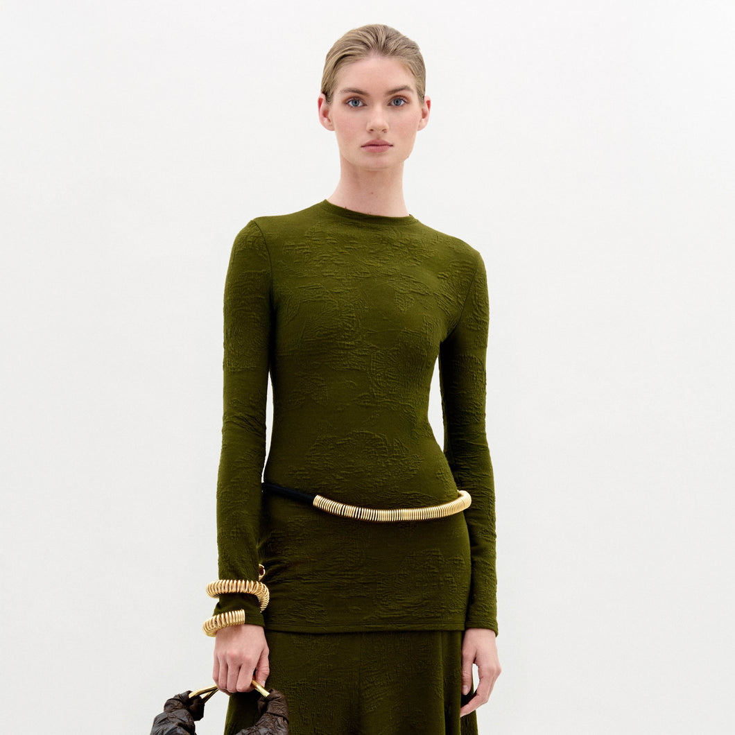 A woman stands against a plain white background wearing the Gen Tshirt, a slim fit, textured olive green long-sleeve dress with gold accessories and holding a brown bag.