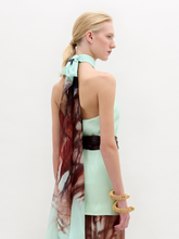 Load image into Gallery viewer, A woman stands against a plain white background wearing the Geovanna Blouse—a sleeveless, high-neck halter top featuring a brown and light blue abstract pattern—paired with a wide belt and matching skirt, made in Colombia.
