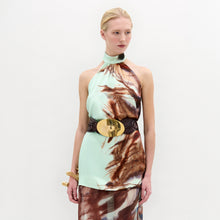 Load image into Gallery viewer, A woman stands against a plain white background wearing the Geovanna Blouse—a sleeveless, high-neck halter top featuring a brown and light blue abstract pattern—paired with a wide belt and matching skirt, made in Colombia.
