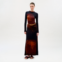 Load image into Gallery viewer, A woman with slicked-back hair and a neutral expression stands against a white background wearing the floor-length, long-sleeved Gabbice Dress in black with bold red floral patterns, styled with a braided belt and dark sandals.
