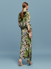 Load image into Gallery viewer, On August 19th, a woman poses against a light blue background wearing the Harriet Dress Floral Camo in green, black, and brown floral pattern. She is accessorized with high-heeled shoes and a bracelet.
