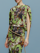 Load image into Gallery viewer, On August 19th, a woman poses against a light blue background wearing the Harriet Dress Floral Camo in green, black, and brown floral pattern. She is accessorized with high-heeled shoes and a bracelet.
