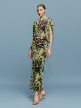 Load image into Gallery viewer, On August 19th, a woman poses against a light blue background wearing the Harriet Dress Floral Camo in green, black, and brown floral pattern. She is accessorized with high-heeled shoes and a bracelet.
