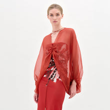 Load image into Gallery viewer, A woman stands against a plain background wearing the Idra Blouse, a sheer, long-sleeved top with a deep V neckline, layered over a patterned shirt and paired with a matching red skirt.
