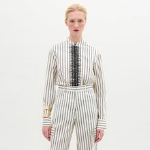 Load image into Gallery viewer, A person stands against a plain background wearing the Isabell Blouse, a white long-sleeve top with black lace and delicate embroidery down the front, paired with pants featuring black vertical stripes.
