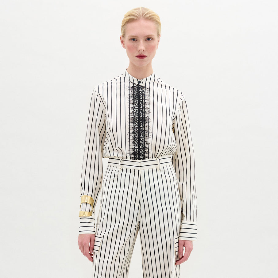 A person stands against a plain background wearing the Isabell Blouse, a white long-sleeve top with black lace and delicate embroidery down the front, paired with pants featuring black vertical stripes.