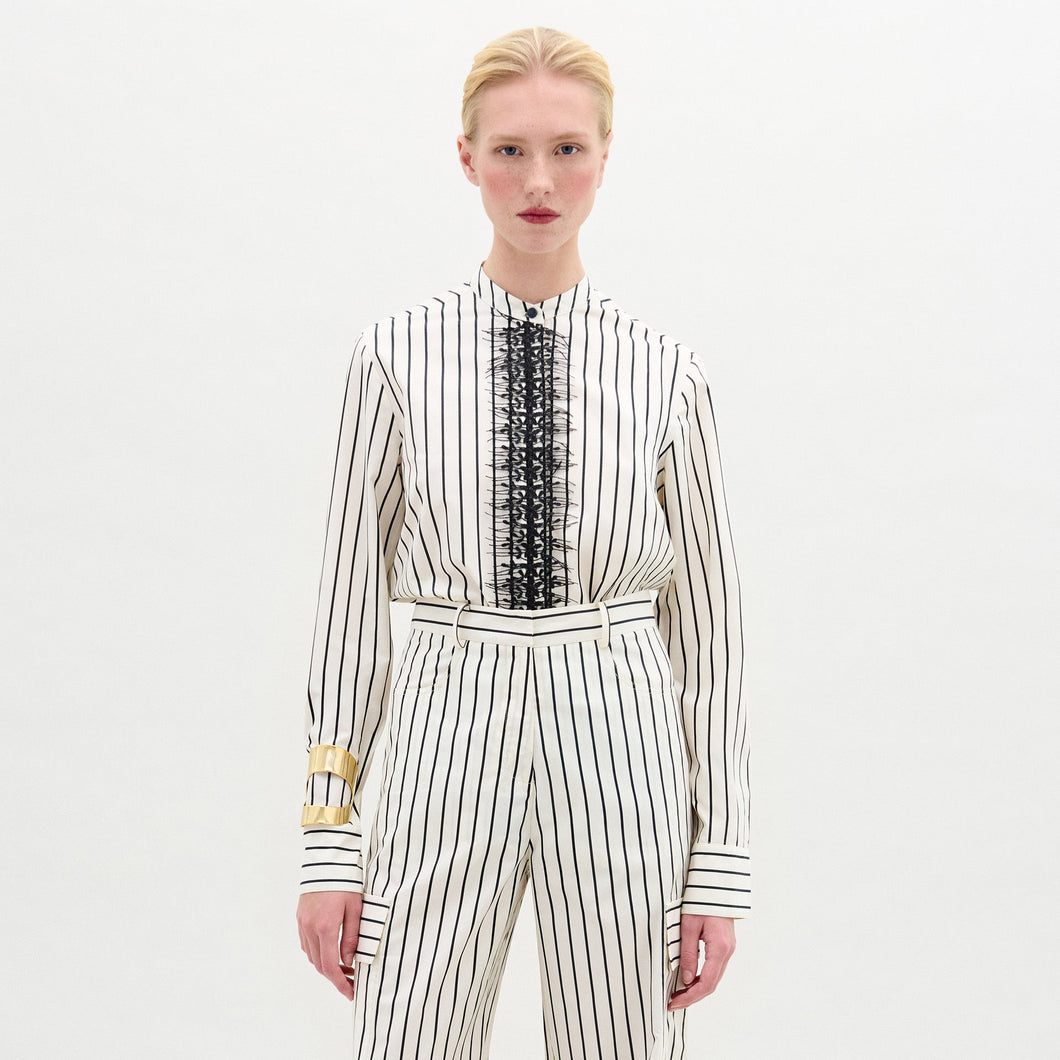 A person stands against a plain background wearing the Isabell Blouse, a white long-sleeve top with black lace and delicate embroidery down the front, paired with pants featuring black vertical stripes.