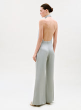 Load image into Gallery viewer, A woman stands against a plain background wearing the Imperio Jumpsuit—a light gray, backless halter style with wide-leg pants and high heels. She is turned to the side, highlighting its open-back design.
