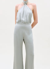 Load image into Gallery viewer, Someone stands against a plain light background wearing the Imperio Jumpsuit, a sleeveless, light gray halter-neck style with wide-leg pants, gathered neckline, and a fitted waist.
