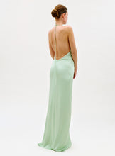 Load image into Gallery viewer, A woman faces away, wearing the Jenifer Dress—a long, light green backless gown with a halter neck tie—her hair in a neat bun against a simple, light background.
