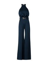 Load image into Gallery viewer, A woman wearing the Kaede Jumpsuit Navy, featuring a halter-neck design with wide-leg pants and a black belt, stands against a light blue background. She accessorizes with multiple black bracelets on one arm, exuding an air of chic nautical elegance.
