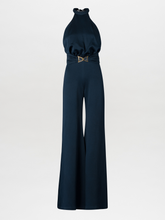 Load image into Gallery viewer, A woman wearing the Kaede Jumpsuit Navy, featuring a halter-neck design with wide-leg pants and a black belt, stands against a light blue background. She accessorizes with multiple black bracelets on one arm, exuding an air of chic nautical elegance.
