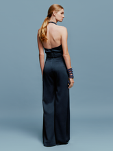 Load image into Gallery viewer, A woman wearing the Kaede Jumpsuit Navy, featuring a halter-neck design with wide-leg pants and a black belt, stands against a light blue background. She accessorizes with multiple black bracelets on one arm, exuding an air of chic nautical elegance.
