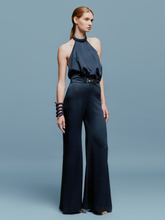 Load image into Gallery viewer, A woman wearing the Kaede Jumpsuit Navy, featuring a halter-neck design with wide-leg pants and a black belt, stands against a light blue background. She accessorizes with multiple black bracelets on one arm, exuding an air of chic nautical elegance.
