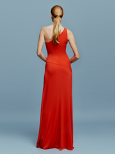 Load image into Gallery viewer, A woman in a Kanya Dress Rouge, featuring a red, one-shoulder design and floor-length cut, stands against a plain blue background. Her hair is pulled back as she calmly stands with her arms by her sides, appearing to wait for an important date such as August 19th or September 2nd.
