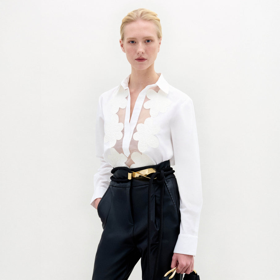 A person with blond hair wears the Karole Blouse, which has long sleeves and floral cutout details, paired with black trousers and a gold belt, standing against a plain white background.