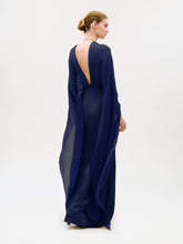 Load image into Gallery viewer, A person stands facing away, wearing the Kalina Dress—a floor-length, deep blue maxi gown crafted from luxurious Italian fabric. The dress features a sheer, flowing design with a low-cut back and column silhouette, while their hair is styled in a neat bun against a plain, light background.
