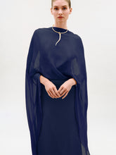 Load image into Gallery viewer, A person wearing the Kalina Dress—a sheer navy blue cape-style maxi crafted from luxurious Italian fabric—paired with a gold choker necklace, stands against a plain light background, looking at the camera with hands gently clasped.
