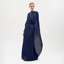 Load image into Gallery viewer, A woman stands in a studio wearing the Kalina Dress—a long, flowing navy blue maxi gown crafted from luxurious Italian fabric, with elegant cape-like sleeves and a gold necklace. The dress’s column silhouette is beautifully highlighted against the plain white background.
