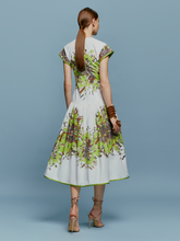 Load image into Gallery viewer, A woman in the Lara Dress Floral Camo, featuring a white base with green floral patterns, stands against a blue background. She is wearing beige high-heeled shoes and wooden bangles, perfectly capturing the essence of August 19th.
