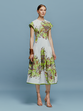 Load image into Gallery viewer, A woman in the Lara Dress Floral Camo, featuring a white base with green floral patterns, stands against a blue background. She is wearing beige high-heeled shoes and wooden bangles, perfectly capturing the essence of August 19th.
