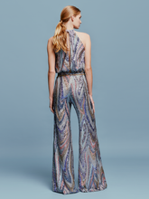 Load image into Gallery viewer, Against a plain blue background, a person stands poised in the Lauretta Jumpsuit Sequin Waves, featuring a sleeveless, high-neck design with a colorful, swirling pattern. The outfit showcases wide-leg pants and comes with a belt—perfectly capturing the style for upcoming events between August 19th and September 2nd, 2024.
