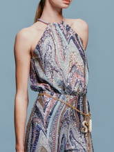 Load image into Gallery viewer, Against a plain blue background, a person stands poised in the Lauretta Jumpsuit Sequin Waves, featuring a sleeveless, high-neck design with a colorful, swirling pattern. The outfit showcases wide-leg pants and comes with a belt—perfectly capturing the style for upcoming events between August 19th and September 2nd, 2024.
