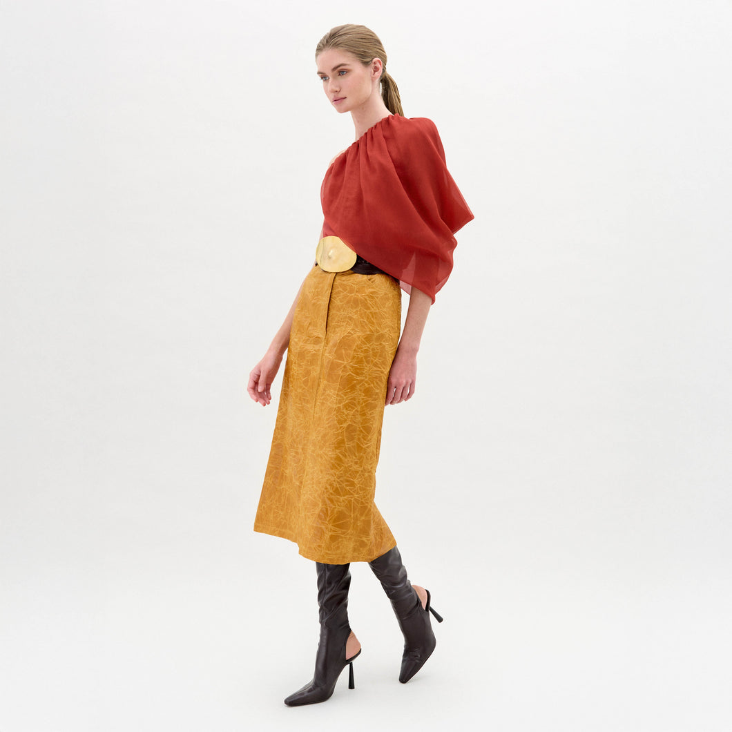 A woman models the Layan Skirt—a mustard yellow structured skirt with a large circular belt—styled with a red blouse and black heeled boots against a plain white background.