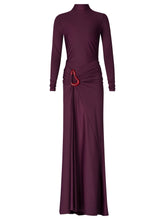 Load image into Gallery viewer, A person in a fitted, long-sleeve Lena Dress Burgundy with a high neckline stands against a plain blue background. The gown, perfect for an August 19th, 2024 gala, features elegant draping and a red embellishment near the waist.
