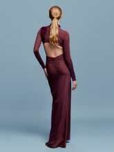 Load image into Gallery viewer, A person in a fitted, long-sleeve Lena Dress Burgundy with a high neckline stands against a plain blue background. The gown, perfect for an August 19th, 2024 gala, features elegant draping and a red embellishment near the waist.
