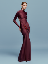 Load image into Gallery viewer, A person in a fitted, long-sleeve Lena Dress Burgundy with a high neckline stands against a plain blue background. The gown, perfect for an August 19th, 2024 gala, features elegant draping and a red embellishment near the waist.
