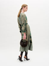 Load image into Gallery viewer, A woman stands against a white background wearing the Erin Skirt—a long, green and beige patterned skirt crafted from Italian fabric—paired with black heeled boots and a small black handbag.
