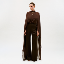 Load image into Gallery viewer, Against a plain white background, a woman wears the Ligia Jumpsuit—a dark brown, cape-like design with wide-leg trousers and sheer fabric draping elegantly from her shoulders to her ankles.
