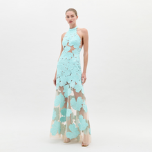 Load image into Gallery viewer, A woman wearing the Mab Dress Turquoise, a light blue floral-patterned halter neck dress with a sheer skirt, stands against a plain background, eagerly awaiting the pre-order ship-by date of February 1st.
